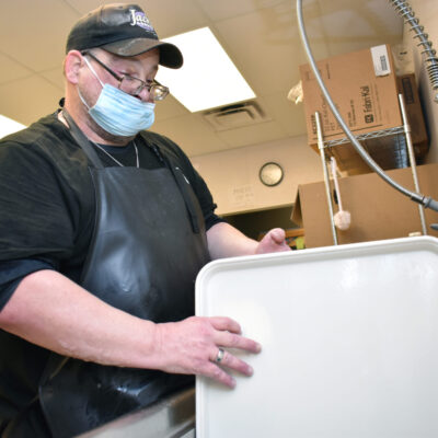 A man with a facemask, glasses and baseball hat washes a large tray