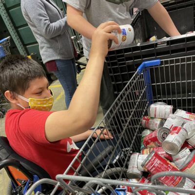 A teen boy in a wheelchair hands canned goods from a cart to another person