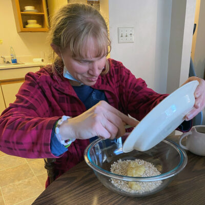 A blonde woman seated at a table scrapes recipes ingredients off a plate into a mixing bowl