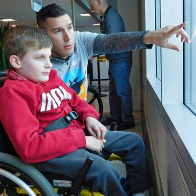 Volunteers-wings A boy sits in a wheellchair looks out a window as a man points to it