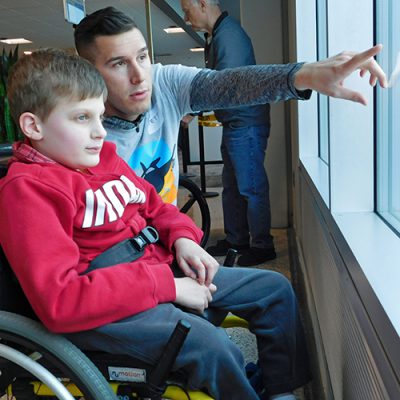 A boy sits in a wheellchair looks out a window as a man points to it