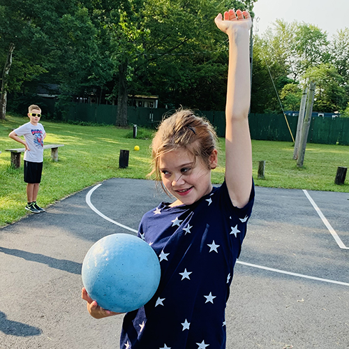 A young girl stands on a playground with one hand in the air and a ball in the other hand