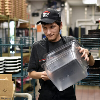 Julian carries plastic boxes to the table where he’s filling small cups with marinara sauce. Those cups of sauce will be stacked in the plastic boxes and stored there until orders are assembled.